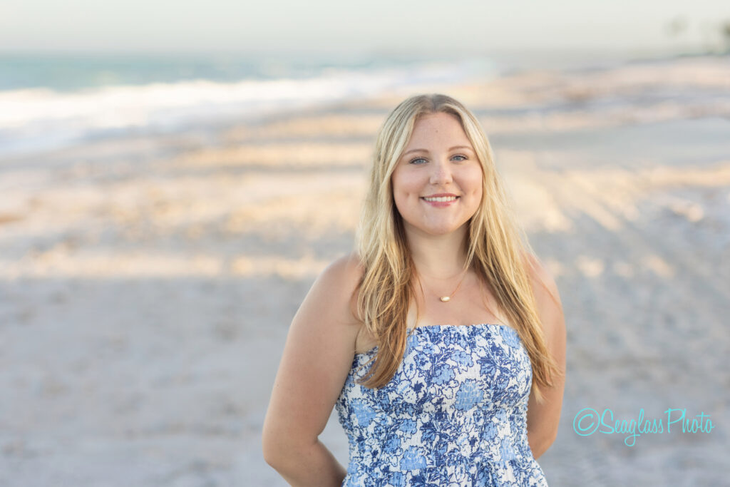 A young woman with long blonde hair, wearing a blue and white floral dress, smiles while standing on a sandy beach with waves in the background under a clear sky. Vero Beach Photoshoot