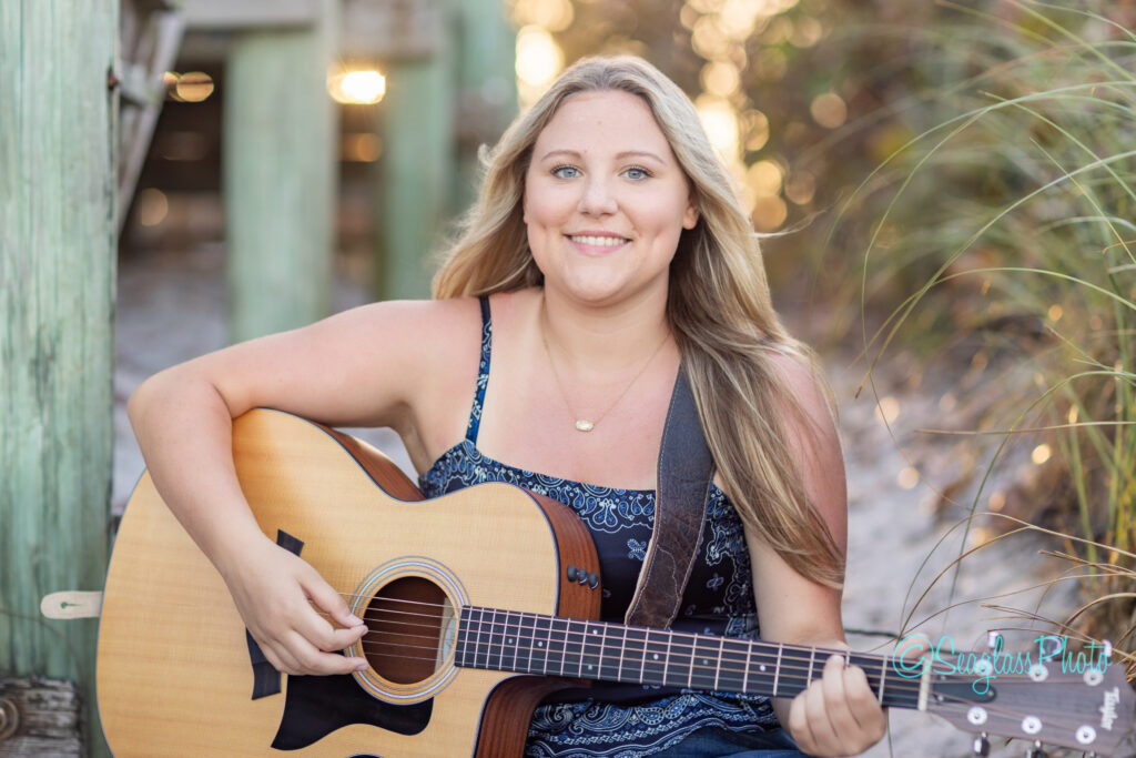 A young woman with long blonde hair, wearing a sleeveless dress, sits outdoors on a sandy path playing an acoustic guitar, smiling at the camera. Sunlight shines through the background with grass and wooden posts visible. Vero Beach Photoshoot