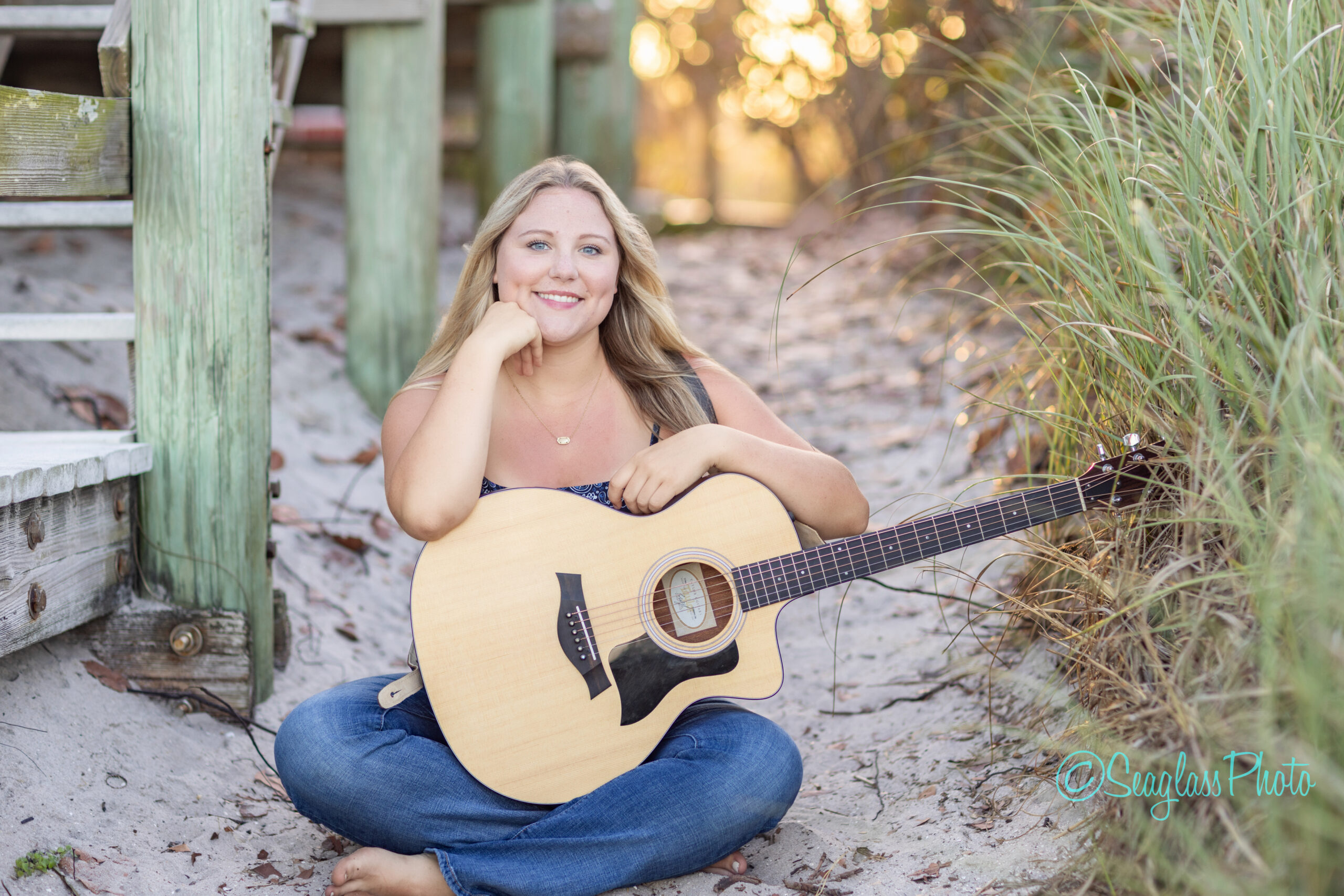 A young woman with long blonde hair sits barefoot on sandy ground near wooden steps, smiling and holding an acoustic guitar, surrounded by beach grass and soft sunlight. Vero Beach Photoshoot