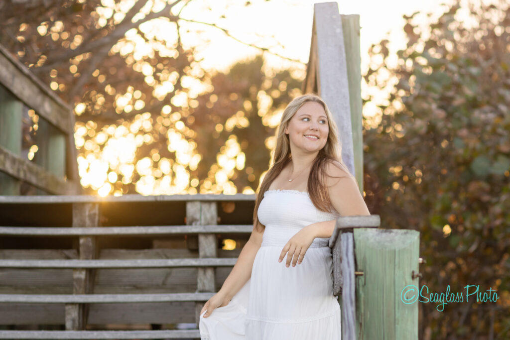 A young woman in a white dress stands on wooden steps outdoors at sunset, smiling and looking to the side, with warm sunlight and autumn foliage in the background. Vero Beach Photoshoot