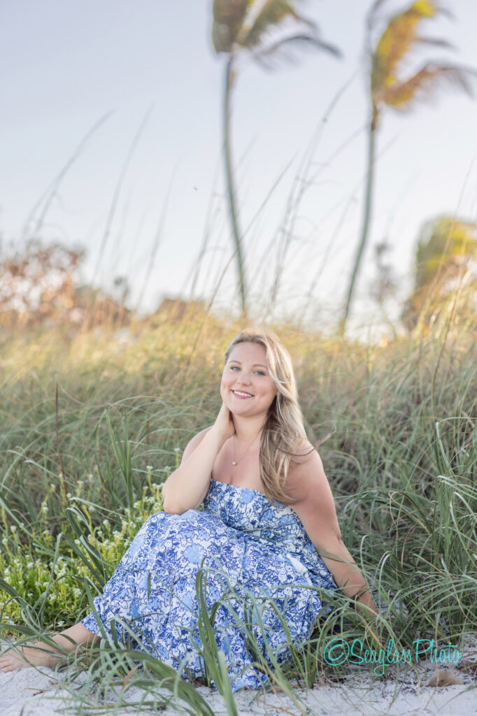 A woman in a blue and white floral dress sits on sandy grass at the beach, smiling with one hand on her neck. Tall grass and palm trees sway in the background under a clear sky. Vero Beach Photoshoot