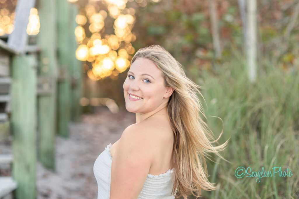 A young woman with long blonde hair, wearing a white off-shoulder top, smiles over her shoulder at the camera. She stands outdoors near a wooden staircase with greenery and soft sunlight in the background. Vero Beach Photoshoot
