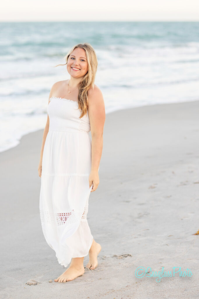 A woman in a white strapless dress walks barefoot along the beach, smiling. The ocean waves and sandy shore are visible in the background under a clear sky. Vero Beach Photoshoot