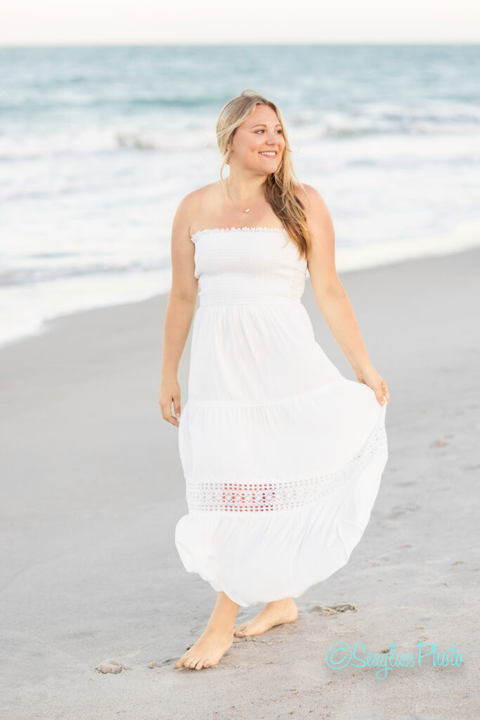 A woman in a white strapless dress walks barefoot along the beach, smiling and looking to the side. The ocean waves and sandy shore are visible in the background. Vero Beach Photoshoot