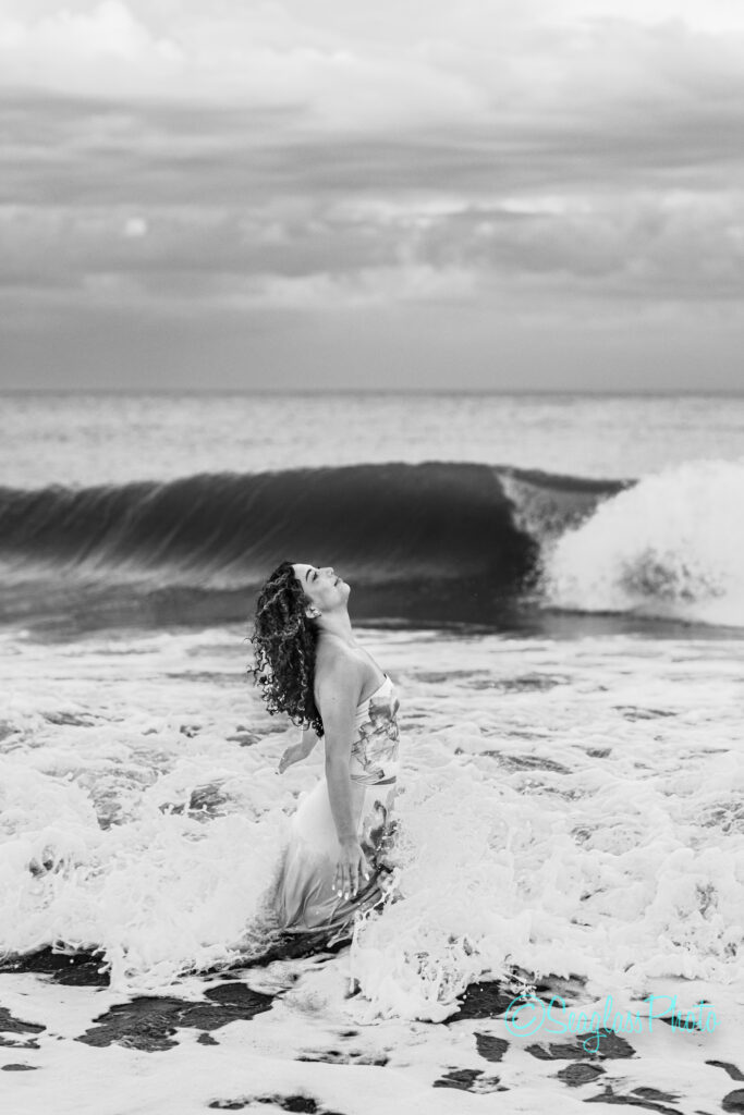 A woman in a long dress kneels in the ocean surf with waves crashing behind her, tilting her head back and eyes closed. The scene is in black and white, under a dramatic cloudy sky. Vero Beach Photoshoot