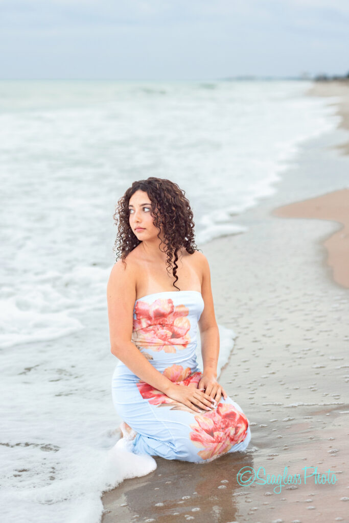 A woman with curly hair wearing a strapless floral dress kneels on wet sand at the edge of the ocean, looking to her left, with gentle waves washing ashore under a cloudy sky. Vero Beach Photoshoot