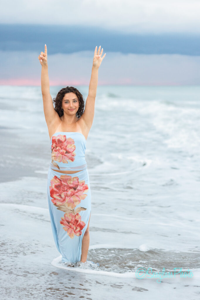 A woman in a light blue dress with large pink flowers stands in shallow ocean water, arms raised, with waves around her feet and a cloudy sky in the background. Vero Beach Photoshoot