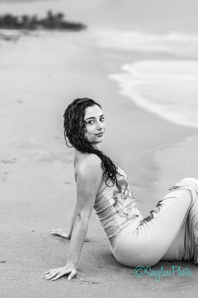 A woman with wet hair sits on the sand at the beach near the shoreline, looking over her shoulder toward the camera. She wears a strapless dress, and the image is in black and white. Vero Beach Photoshoot