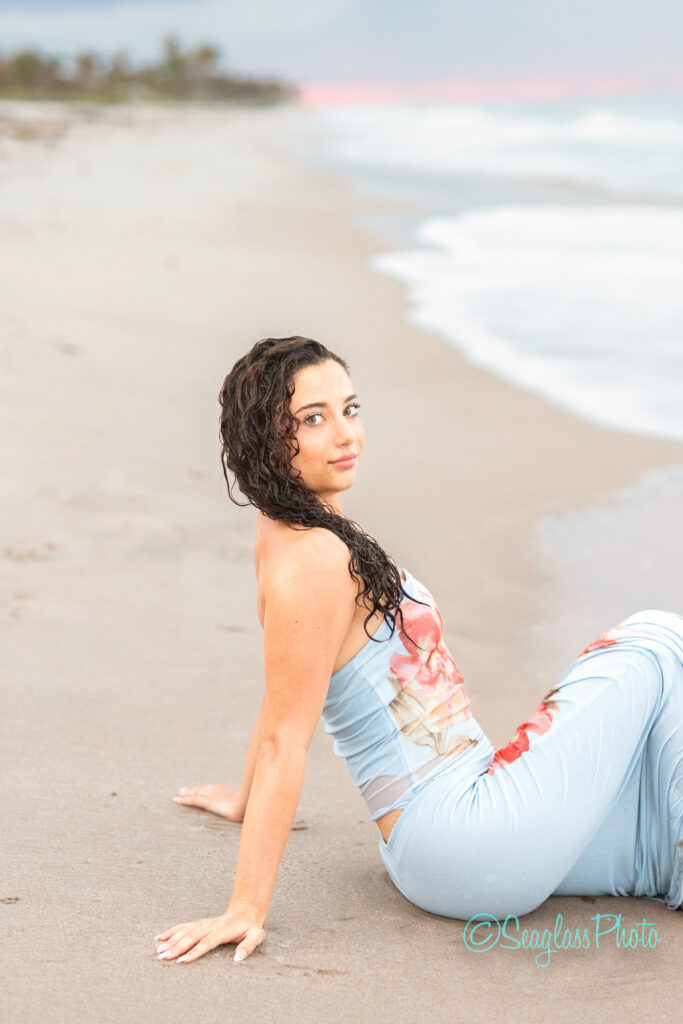 A woman with wet, curly hair sits on a sandy beach near the water, wearing a light blue floral dress and looking over her shoulder. The ocean and sky appear soft and muted in the background. Vero Beach Photoshoot