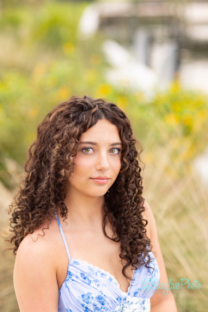 A young woman with long, curly brown hair wearing a blue floral dress looks at the camera with a soft expression. She is outdoors, with blurred greenery and yellow flowers in the background. Vero Beach Photoshoot