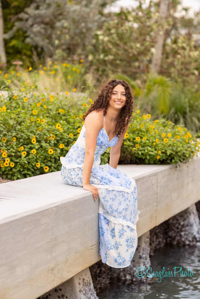 A woman with curly hair, wearing a long blue and white floral dress, sits on a concrete ledge and smiles. Yellow flowers and greenery are in the background on a cloudy day. Vero Beach Photoshoot