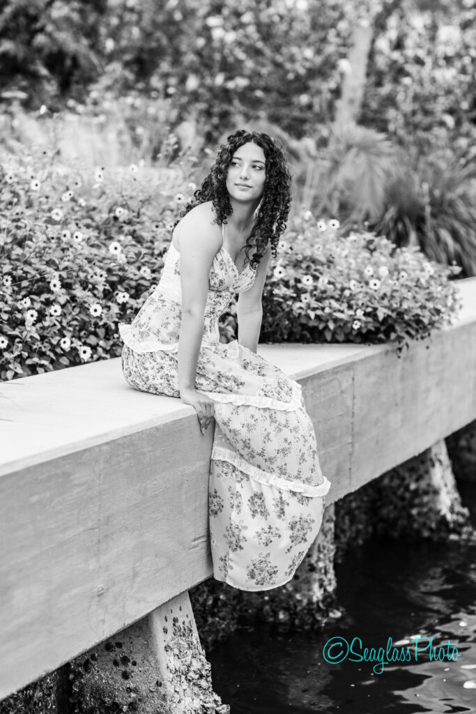 A young woman with curly hair in a floral dress sits on a wooden ledge by a flower garden, with her legs hanging over the edge above water. The image is black and white, with Seaglass Photo logo in the corner. Vero Beach Photoshoot