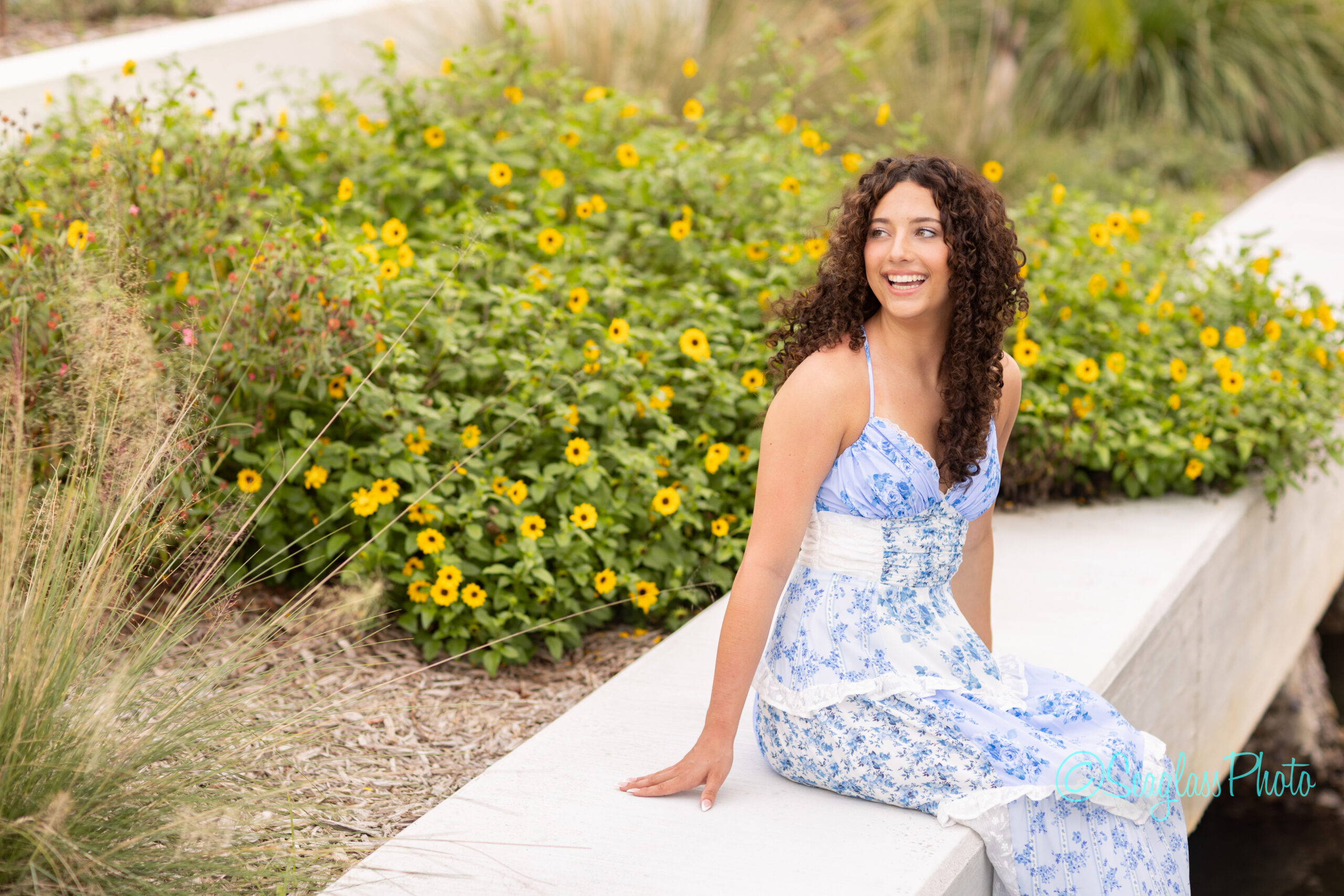 A young woman with curly hair in a blue and white floral dress sits on a concrete bench, smiling and looking to the side, with blooming yellow flowers and greenery in the background. Vero Beach Photoshoot