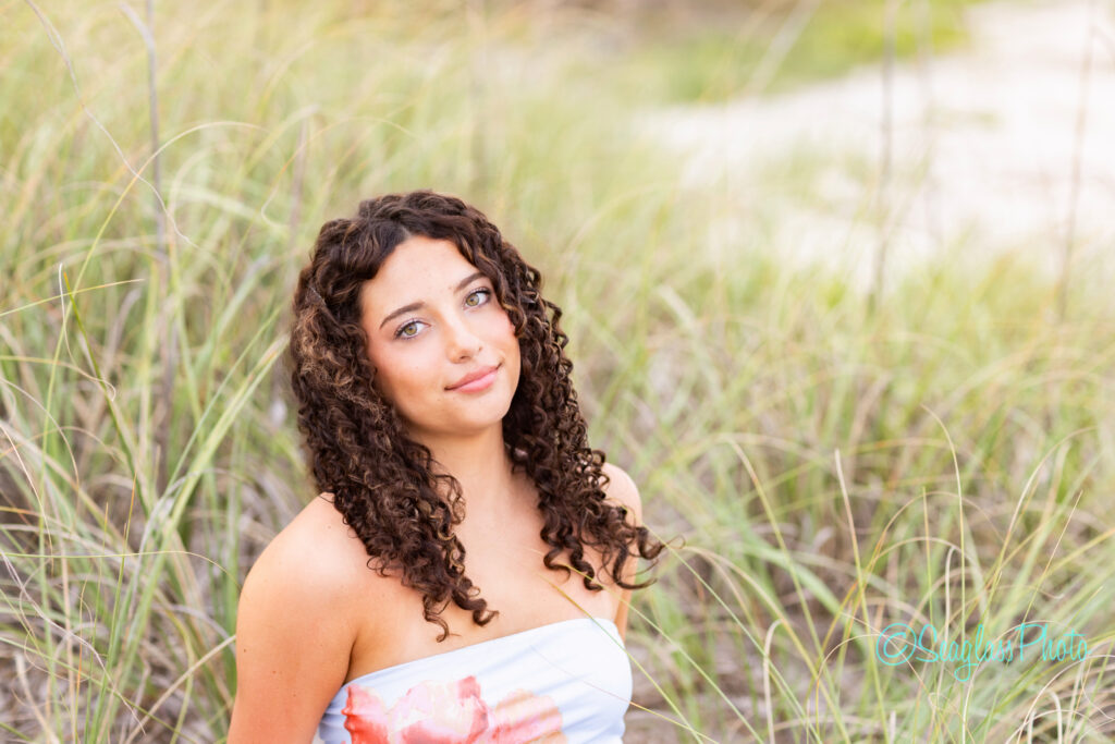 A young woman with curly brown hair and blue eyes stands in front of tall grass, wearing a strapless dress and looking slightly upward with a gentle expression. The background is softly blurred. Vero Beach Photoshoot