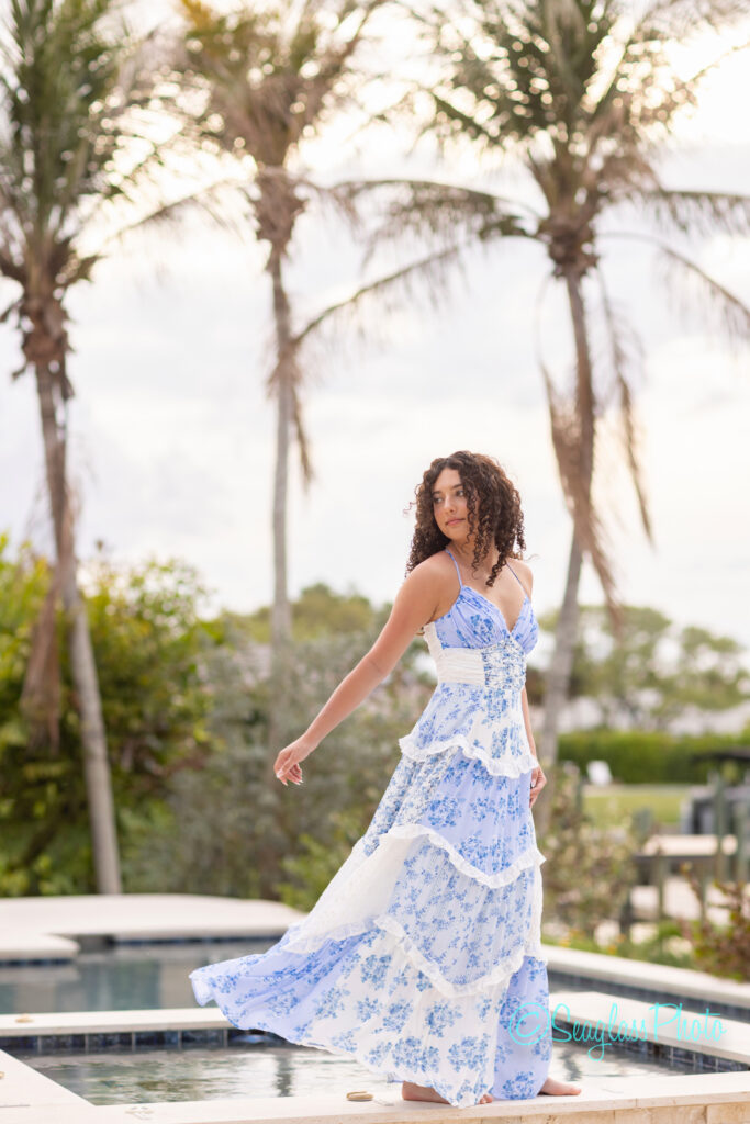 A woman in a flowing white and blue floral dress stands near a pool, turning slightly to look over her shoulder. Palm trees and greenery are visible in the background under a cloudy sky. Vero Beach Photoshoot