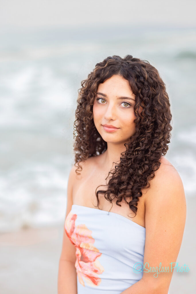 A young woman with long, curly brown hair stands on a beach, wearing a light blue strapless dress with a pink floral design. The ocean waves and sky are softly blurred in the background. Vero Beach Photoshoot