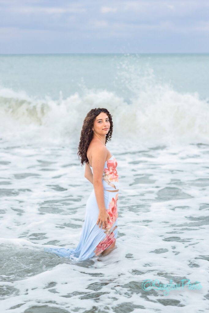 A woman with curly hair in a floral strapless dress stands in the ocean, partially submerged in the water, with waves crashing behind her under a cloudy sky. Vero Beach Photoshoot