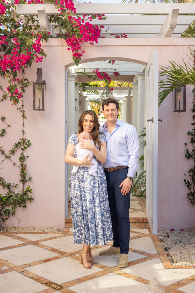 A smiling couple stands outdoors in front of a pink wall and open white gate, holding a baby. The woman wears a blue and white dress; the man wears a light blue shirt and dark pants. Pink flowers and greenery surround them. Vero Beach Photoshoot