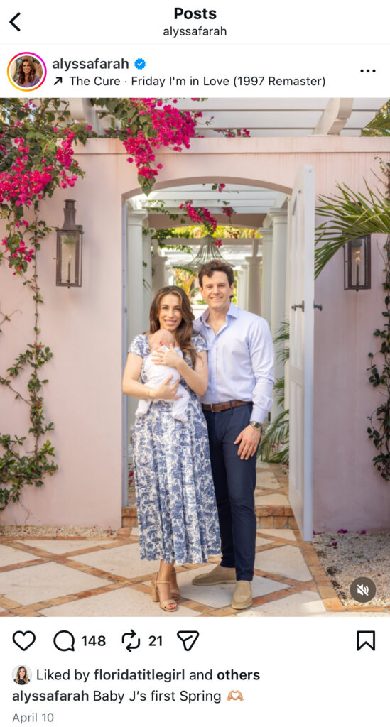 A woman and man stand smiling in a garden walkway, holding a baby. The woman wears a blue-patterned dress, the man wears a white shirt and blue pants. Pink flowers and greenery decorate the background. Vero Beach Photoshoot