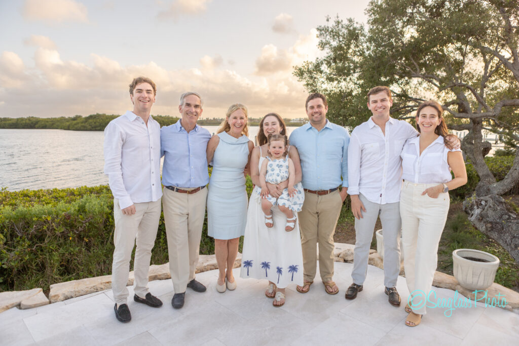 Eight people, including a young child, stand outdoors on a stone patio by the water, smiling for a group photo at sunset. They are dressed in light, summery clothing, with trees and sky in the background. Vero Beach Photoshoot