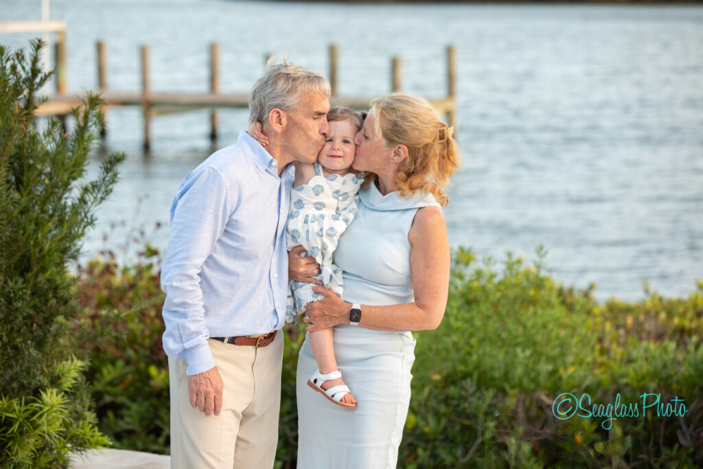 An older man and woman kiss a young child on the cheeks while standing outdoors near water, with greenery and a dock in the background. All three are dressed in light-colored clothes. Vero Beach Photoshoot