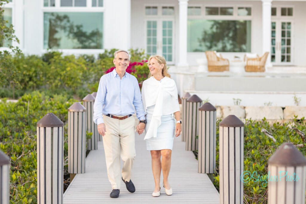 An older couple is walking hand in hand on a wooden pathway, smiling at each other, with a white house and patio furniture in the background surrounded by greenery. Vero Beach Photoshoot