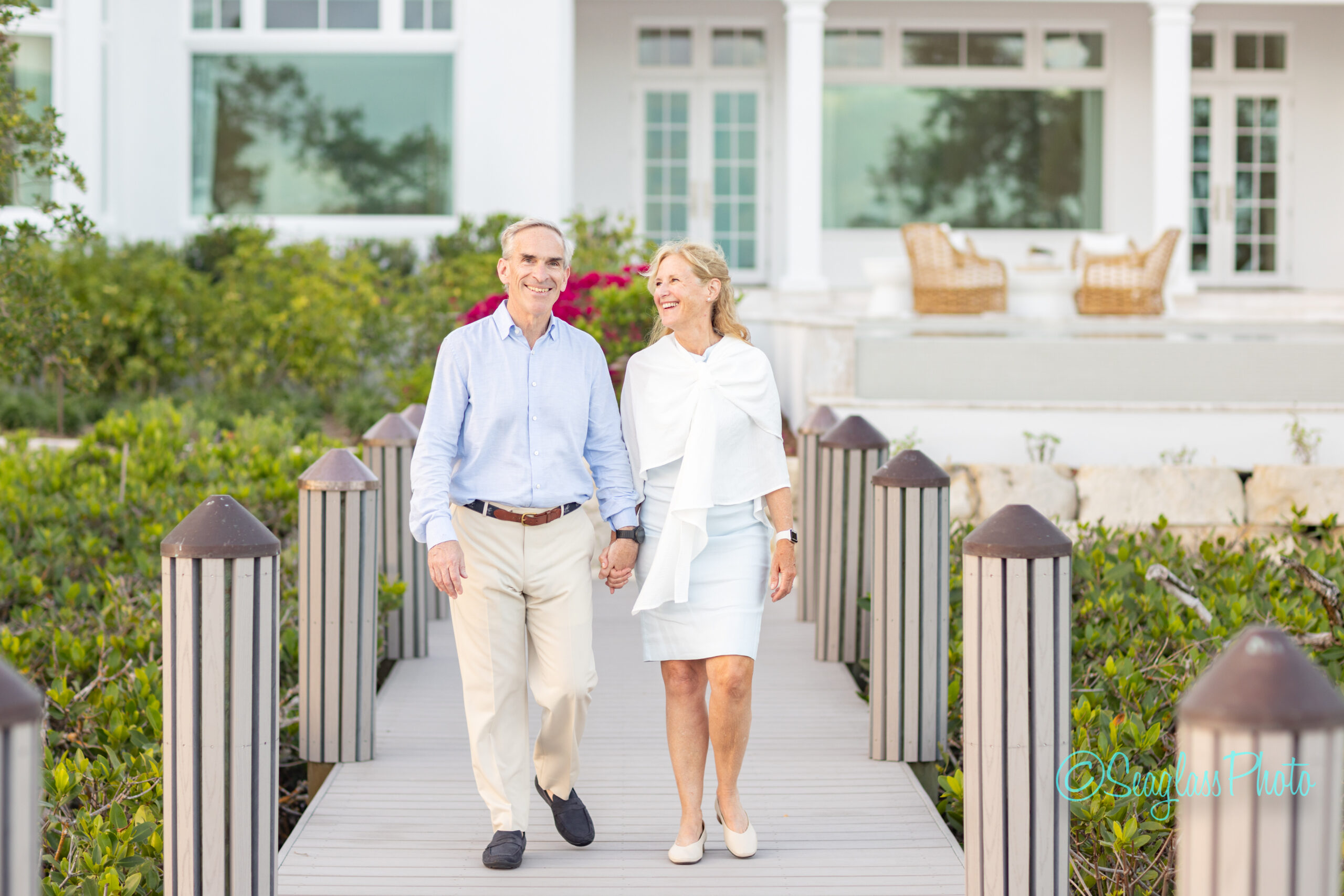 An older couple is walking hand in hand on a wooden pathway, smiling at each other, with a white house and patio furniture in the background surrounded by greenery. Vero Beach Photoshoot