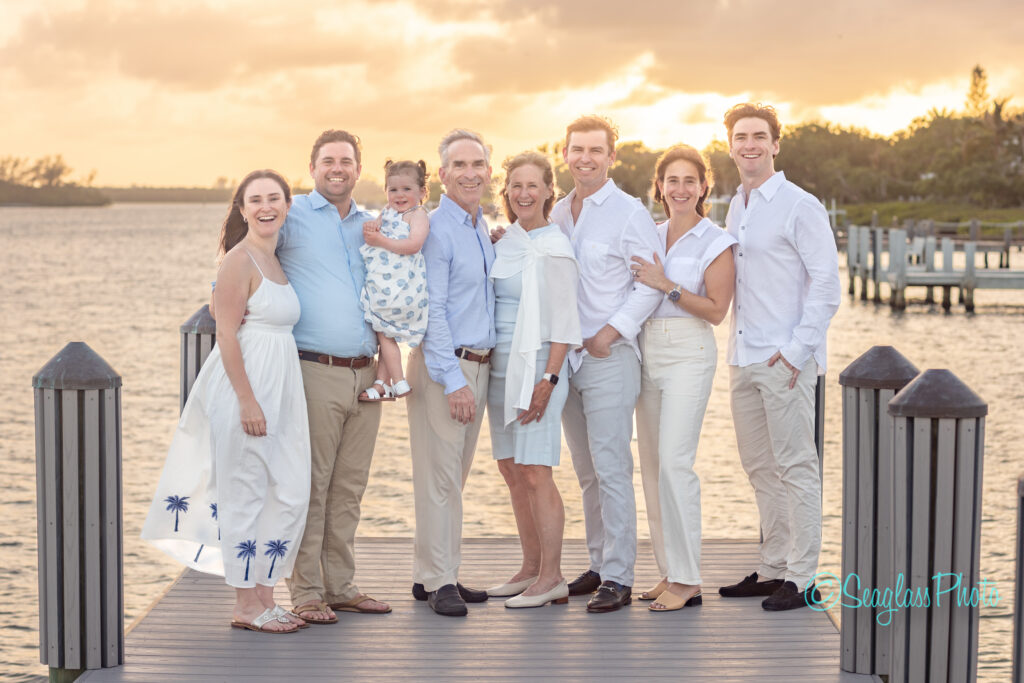 Eight people, including one child, stand close together on a dock by the water at sunset. They are dressed in coordinated white and pastel outfits, smiling and embracing each other with a scenic waterfront and pier in the background. Vero Beach Photoshoot