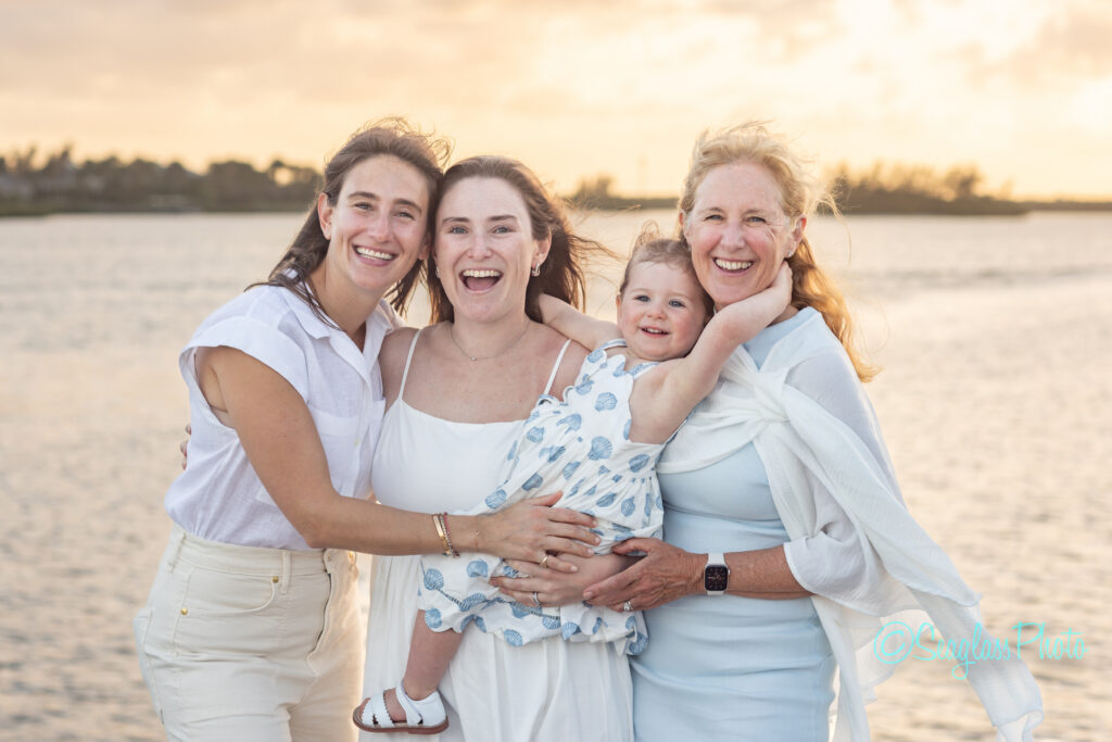 Four smiling women and a young child stand together outdoors by the water at sunset, dressed in light-colored clothing, with one woman holding the child as they all embrace, enjoying a joyful moment. Vero Beach Photoshoot