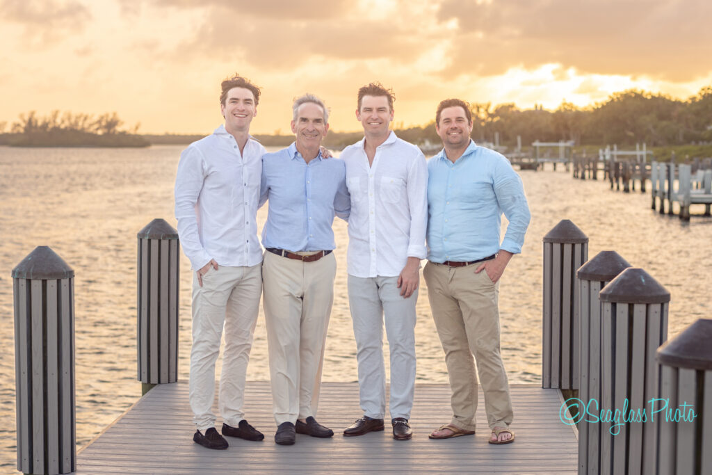 Four men stand smiling on a dock by the water at sunset, wearing light-colored, casual clothes. The sky is golden with clouds, and pilings line the dock. Trees and more docks are visible in the background. Vero Beach Photoshoot