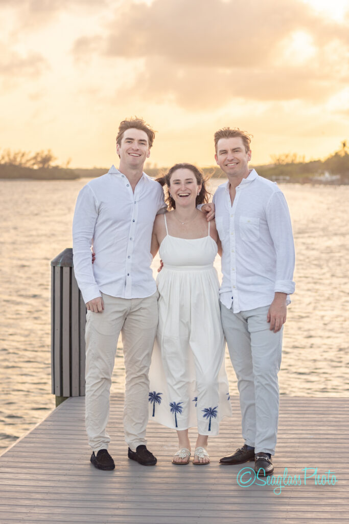 Three people stand on a dock by the water at sunset, smiling. Two men in white shirts and tan pants stand on either side of a woman in a white dress with blue palm trees. The scene is warm and relaxed. Vero Beach Photoshoot