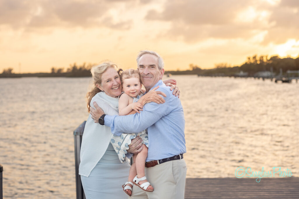An older couple stands on a dock by the water at sunset, smiling and hugging a young child between them. The sky is golden and trees are visible in the distance. Vero Beach Photoshoot