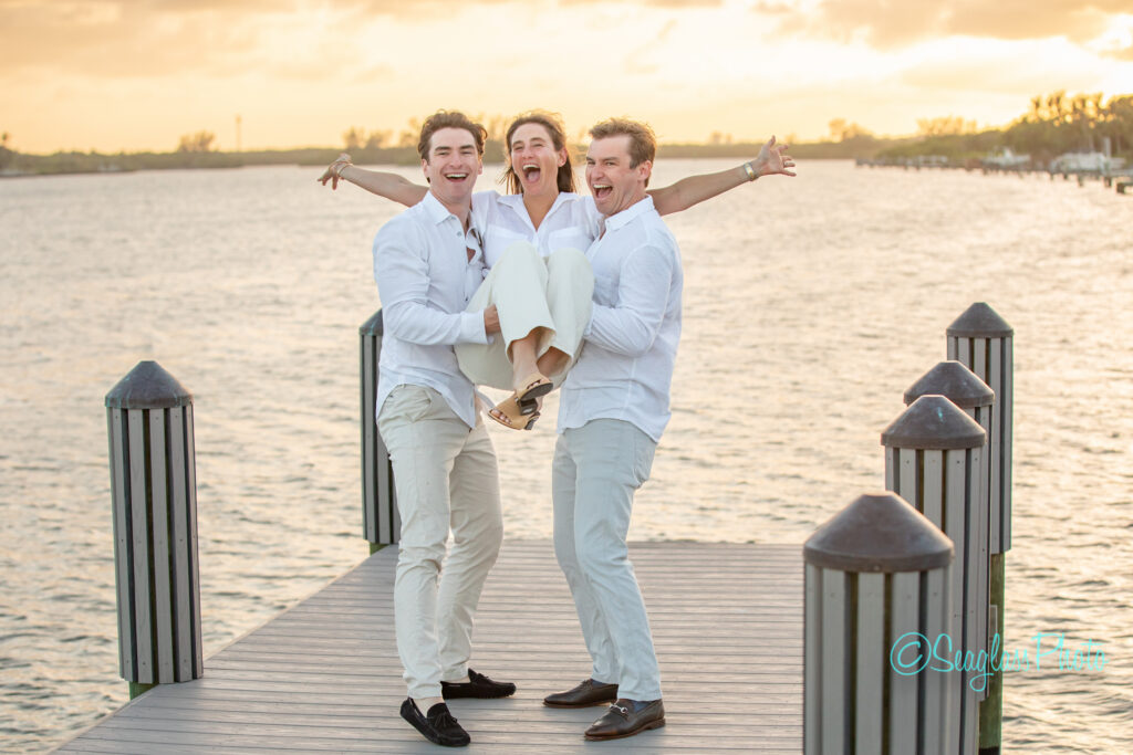 Three people dressed in white stand on a wooden dock by the water at sunset. Two men lift and hold a smiling woman between them, all laughing and appearing joyful. Vero Beach Photoshoot