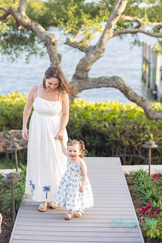 A woman in a white dress walks hand-in-hand with a smiling young girl in a white patterned dress on a wooden path, surrounded by greenery, flowers, and a large tree near water. Vero Beach Photoshoot