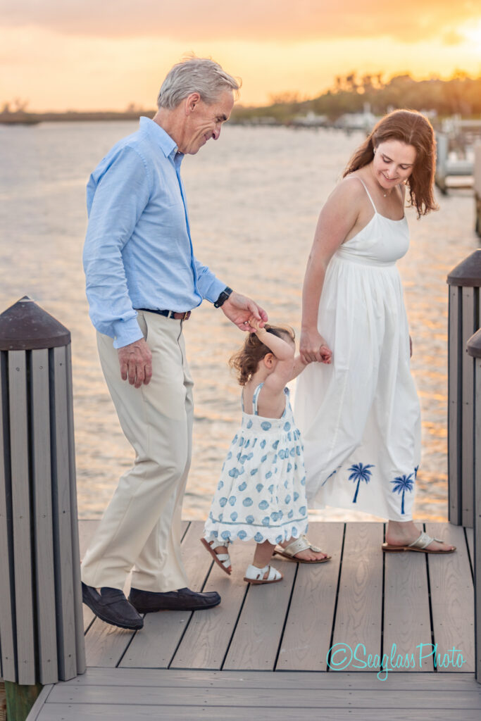 A smiling man, woman, and young girl in summer clothes walk hand-in-hand on a dock by the water at sunset. The little girl wears a white dress with blue designs and holds the womans hand. Vero Beach Photoshoot