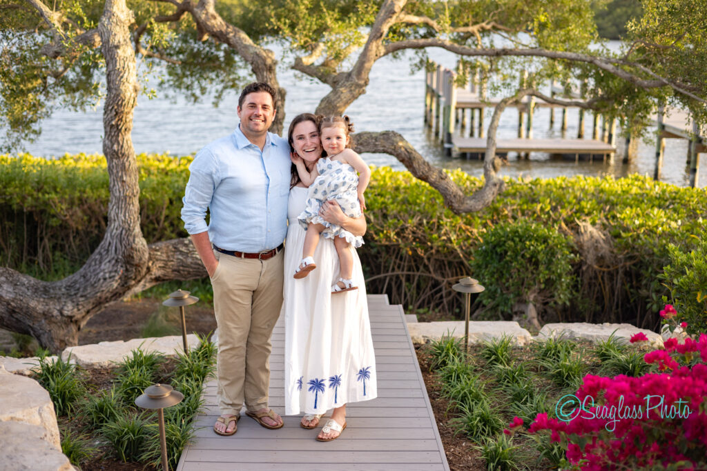 A smiling family of three stands on a wooden path near lush greenery and water. The father wears a light blue shirt and khakis, the mother in a white dress holds their small child. Trees and a dock are visible in the background. Vero Beach Photoshoot