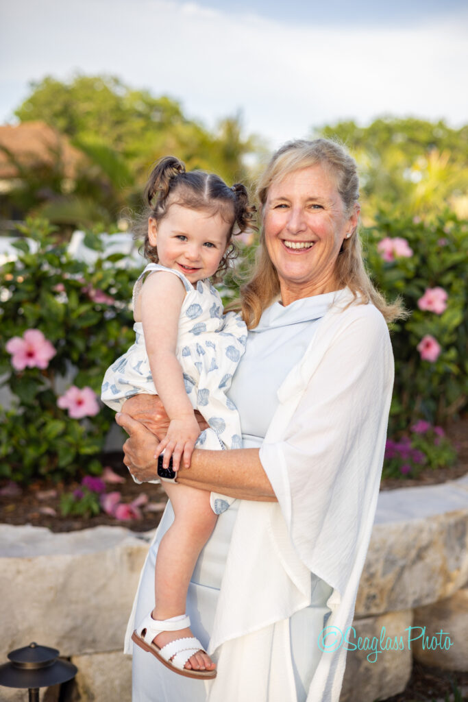 A smiling woman holds a young girl with pigtails outdoors, standing in front of blooming pink flowers and greenery, both wearing light-colored summer outfits. Vero Beach Photoshoot