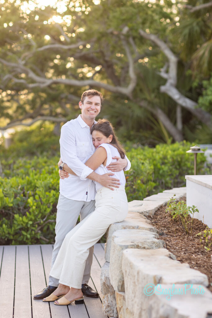 A smiling man in a white shirt hugs a woman in white pants and a white top, who sits on a stone wall. They are surrounded by greenery and sunlight in an outdoor setting. Vero Beach Photoshoot
