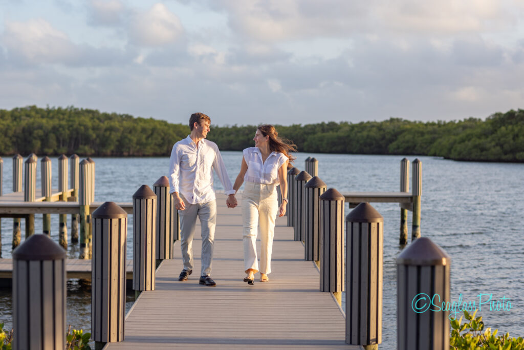 A man and a woman holding hands and smiling at each other as they walk on a wooden dock over the water, surrounded by posts and greenery under a partly cloudy sky. Vero Beach Photoshoot