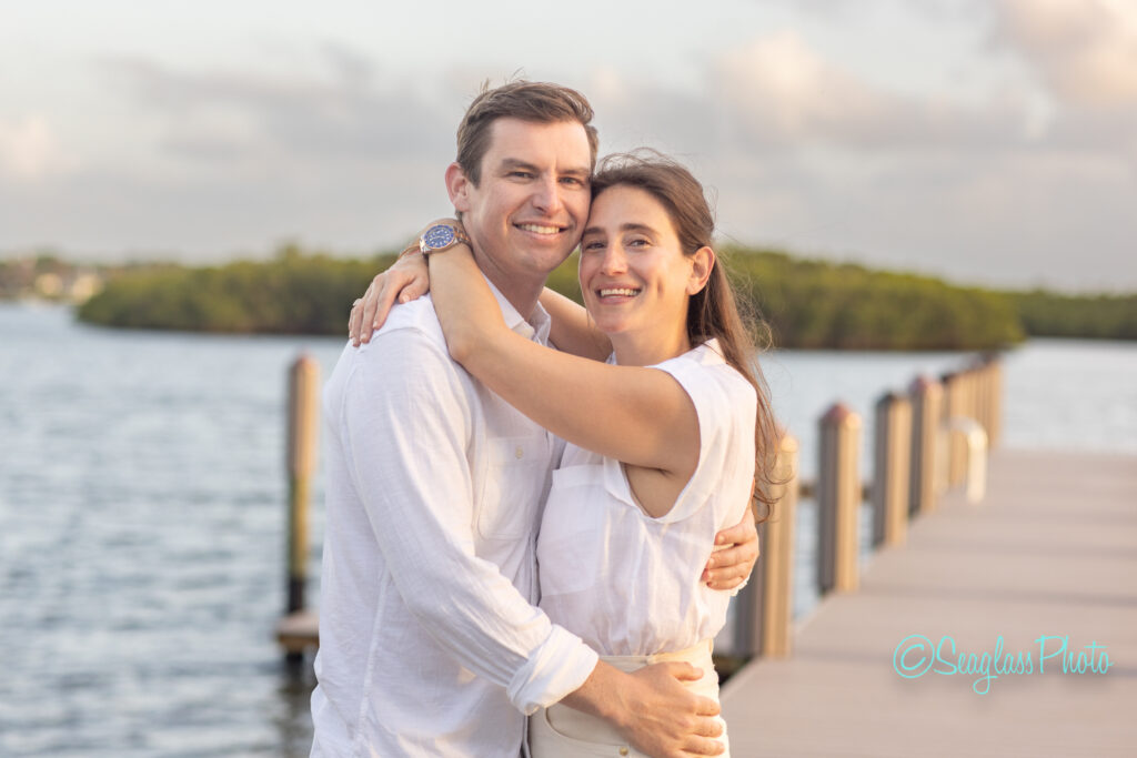 A smiling couple in white clothing embraces on a dock over the water, with green trees and a cloudy sky in the background. Vero Beach Photoshoot