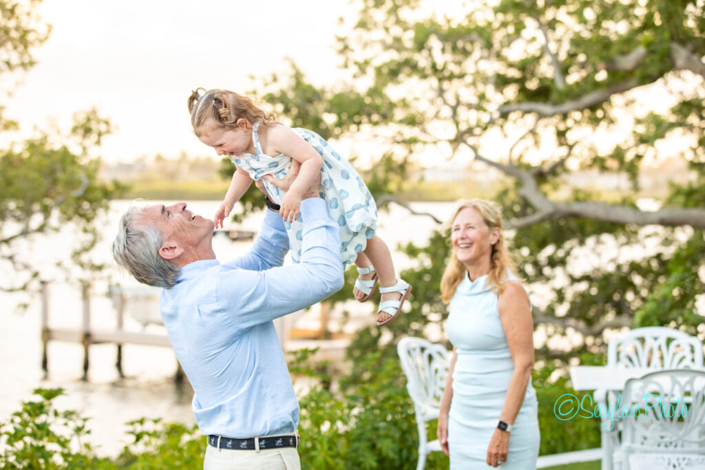 A smiling older man lifts a laughing young girl in a blue polka dot dress above his head outdoors, while a woman in a light blue dress stands nearby, also smiling. Trees and a dock by the water are in the background. Vero Beach Photoshoot