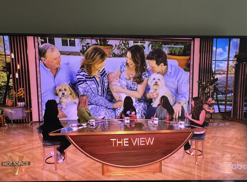 Five women on the set of The View face a large screen displaying a family photo of five adults, a baby, and two dogs, all smiling and sitting closely together outdoors. Vero Beach Photoshoot