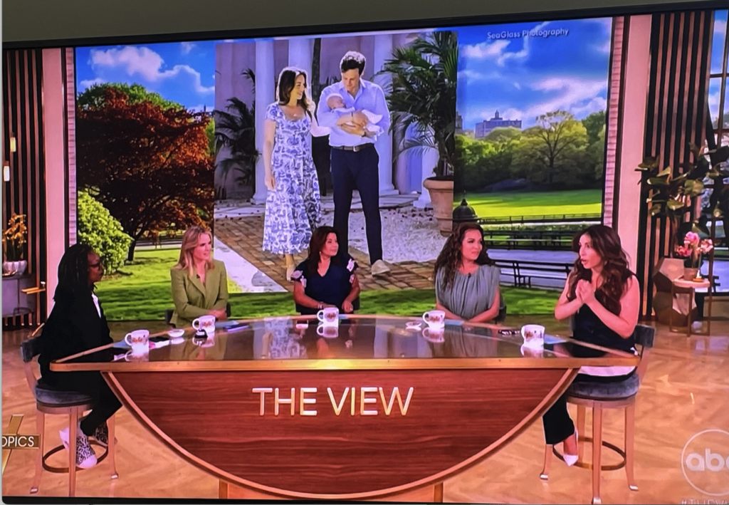 Five women sit at a round table on the set of The View, with a large screen behind them displaying an image of a couple holding a baby outdoors. The ABC logo is visible in the bottom right corner. Vero Beach Photoshoot