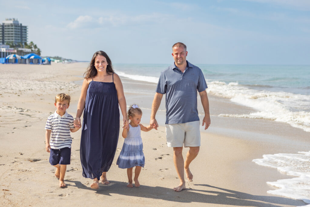 A family of four walks barefoot along a sunny beach. The parents hold hands with their young son and daughter. Waves are gently rolling onto the shore, and buildings and blue umbrellas are visible in the background. Vero Beach Photoshoot