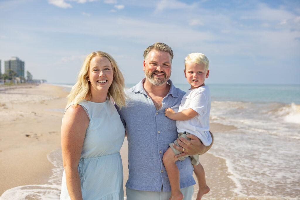 A smiling family of three—woman, man holding a young boy—stands on a sandy beach by the ocean under a blue sky with scattered clouds. Buildings and palm trees are visible in the background. Vero Beach Photoshoot