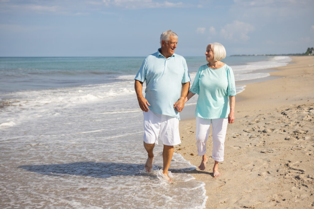 An older couple wearing light blue tops and white shorts walks barefoot along a sandy beach, holding hands and smiling at each other, with gentle waves and a clear sky in the background. Vero Beach Photoshoot
