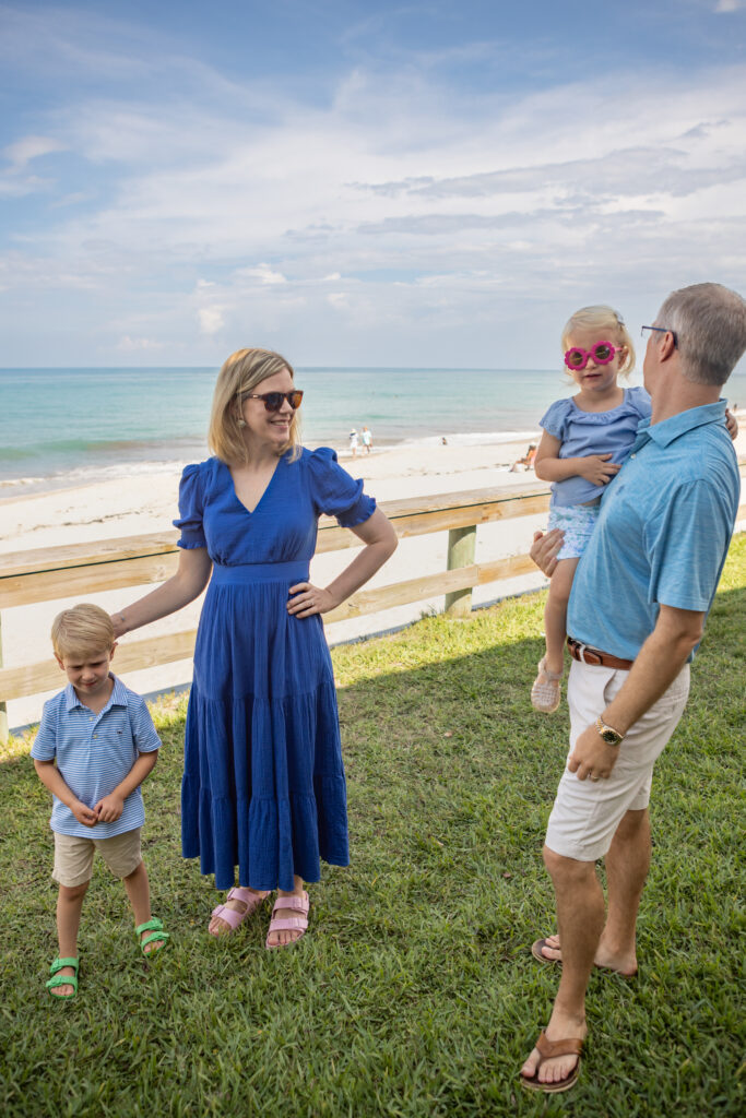 A family of four stands on grass by the beach. The woman in a blue dress smiles, the man holds a girl in pink sunglasses, and a boy stands nearby. The ocean and blue sky are in the background. Vero Beach Photoshoot