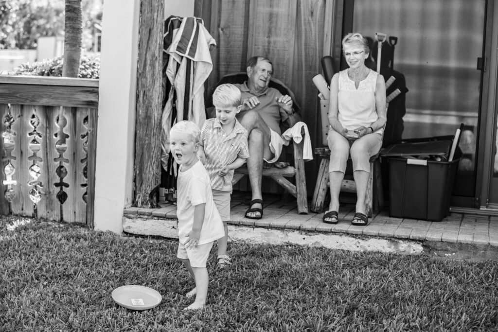 Two young boys play with a frisbee on grass while an older couple smiles, seated on a porch behind them. The scene is outdoors, relaxed, and in black and white. Vero Beach Photoshoot