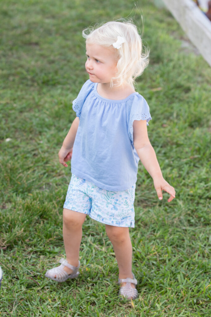 Young child with blonde hair in a light blue shirt, patterned shorts, and clear sandals stands on grass, looking to the side with a slight smile. Vero Beach Photoshoot
