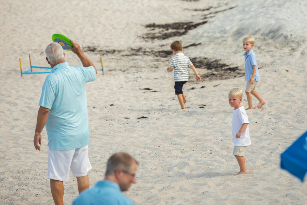 An older man in a light blue shirt holds up a green frisbee on a sandy beach, while four young boys play nearby. One man sits in the foreground, slightly out of focus. Vero Beach Photoshoot